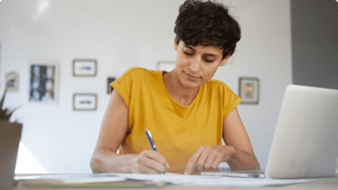 A woman with short dark hair writing on paperwork next to a laptop.